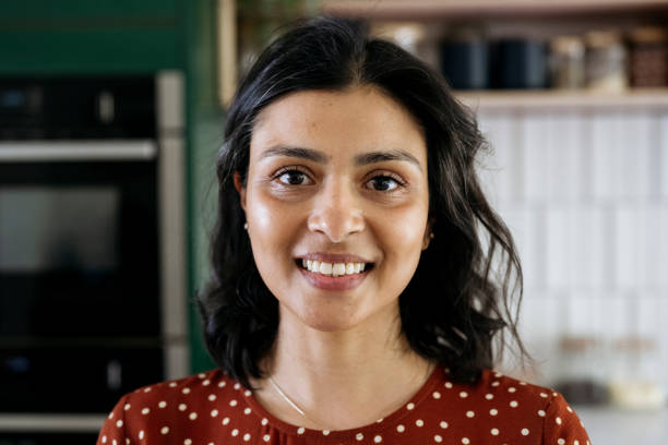 Headshot of early 40s adult with shoulder length black hair, wearing polka dot dress, standing in kitchen, smiling at camera.